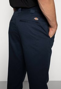 Gros plan sur un pantalon Dickies bleu marine avec poches arrière, passants de ceinture, et une main dans la poche avant droite.