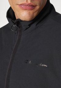 Chaqueta negra hecha de tela suave que presenta un cuello alto, cremallera de longitud completa y un logo reflectante de "Champion" en el pecho.