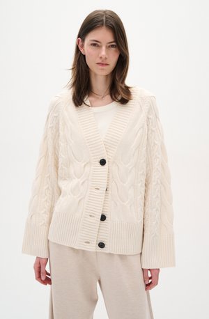 Cremfarbener, gestrickter Cardigan mit Zopfmuster, gerippten Bündchen und Saum sowie schwarzen Knöpfen, über einem hellen Oberteil getragen.