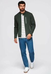 Mörkgrön zip-up jacka med ribbad textur, bärs över en vit och svart randig skjorta, i kombination med blå jeans och vita sneakers.