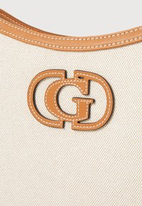 Tissu beige texturé avec des détails en cuir brun, affichant un logo double "G" découpé. Des coutures blanches accentuent les contours du logo.