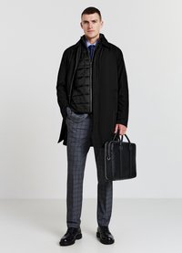 Homme portant un manteau et une veste noirs en couches, une cravate bleue, un pantalon à carreaux gris, des chaussures noires, tenant une mallette noire sur fond blanc.