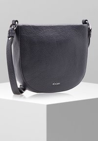 Czarna torebka crossbody z teksturowanej skóry z srebrnymi akcesoriami i logo "JOOP!", spoczywająca na białym pedestale na szarym tle.