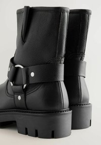 Black leather ankle boots feature a textured finish, a wide strap with a silver ring accent, and a chunky rubber sole with grip patterns.