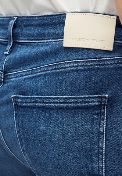 Σκούρο μπλε τζιν με υφή στην επιφάνεια, με λευκή δερμάτινη ετικέτα ραμμένη στη ζώνη μέσης, που εμφανίζει το όνομα της μάρκας.