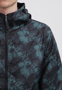 Kurtka z kapturem w czarno-turkusowy wzór tie-dye, wykonana z lekkiego materiału, z centralnym zamkiem błyskawicznym i regulowanym sznurkiem przy kapturze.