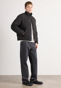 Young man standing, wearing a black quilted zip-up jacket, white shirt, dark loose jeans, and black leather shoes against a plain wall.