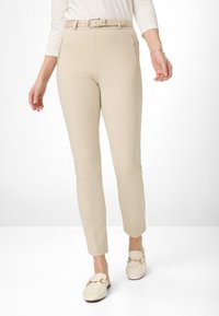 Pantalons beige fuselés avec une ceinture assortie, comportant deux poches avant zippées et une texture de tissu lisse. Portés avec des mocassins de couleur crème.