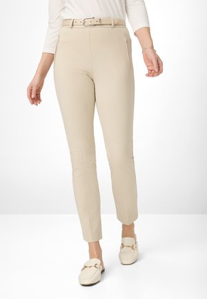 Beige enganzt geschnittene Hose mit einem passenden Gürtel, ausgestattet mit zwei Reißverschlusstaschen vorne und einer glatten Stoffstruktur. Getragen mit cremefarbenen Slippern.