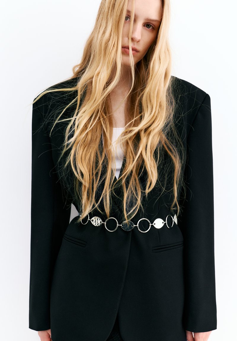 Femme aux longs cheveux blonds portant un blazer noir aux épaules structurées et une ceinture argentée décorative à la taille.