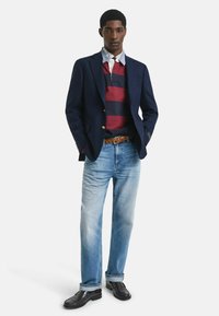 Junger Mann in dunkelblauem Blazer, rot-navy gestreiftem Rugby-Shirt, hellblauen Jeans, braunem geflochtenem Gürtel und dunklen Mokassins, steht mit den Händen in den Taschen.