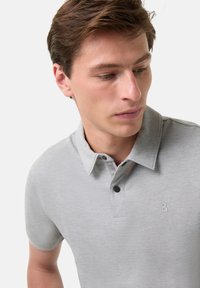 Světlé šedé polo tričko s límečkem, dvěma knoflíky a malým logem značky na levé hrudi. Měkký, hladký textilní materiál.