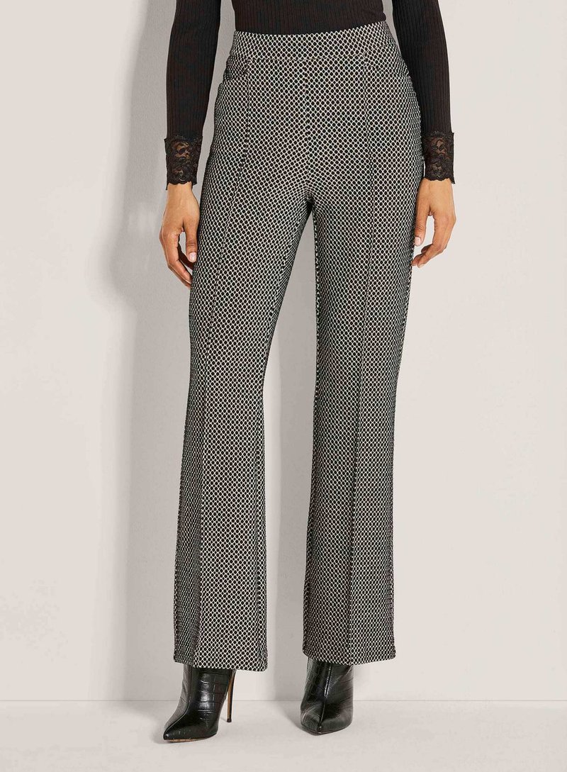 Vrouw in een zwart-wit gestreepte broek met hoge taille en wijde pijpen, met zwart puntig hakken enkellaarzen en een zwart shirt met kant in de mouwen.