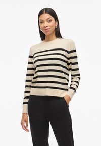 Beige sweater with black stripes, round neckline, long sleeves, and a relaxed fit. Worn over black trousers. Soft knit texture.