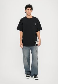 Svart oversized t-skjorte med trykt tekst, kombinert med lyseblå vide jeans og svart-hvite sneakers. Tilbehøret inkluderer en sølvveske.