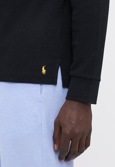 Camisa de manga longa em malha waffle preta com o logotipo dourado da polo na bainha; combinada com calças azul claro. Apresenta fendas laterais e punhos em contraste.