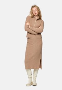 Beige Zweiteiler, bestehend aus einem kurz geschnittenen, mit Reißverschluss versehenen Pullover und einem Midi-Rock mit einem seitlichen Schlitz, aus strukturiertem Stoff. Weiße kniehohe Stiefel.