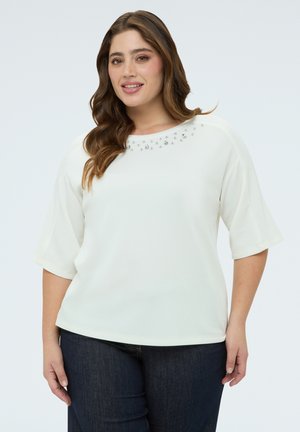 Frau mit langem, welligem braunem Haar, die ein weißes Oberteil mit Strassverzierungen in der Nähe des Halsausschnitts und dunkelblaue Jeans trägt.