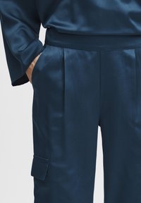 Pantaloni din satin albastru marin cu o textură netedă, dotate cu plii frontale și o buzunar lateral cu clapetă. Croială ajustată pe șolduri.