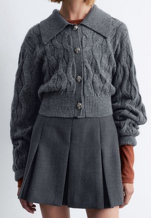 Grå kabelstrikket cardigan med sølv roseknapper over rustfarvede ærmer, kombineret med et mørkegråt plisseret skørt.