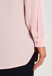 Blusa rosa chiaro con maniche lunghe, tessuto morbido, semplice dettaglio dei polsini con bottone e orlo arrotondato, abbinata a un tessuto scuro sotto.