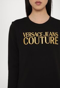 Melnā sporta krekla ar zeltītu izšuvumu "VERSACE JEANS COUTURE" uz krūtīm. Ribu kakls, garas piedurknes, gluda tekstūra.