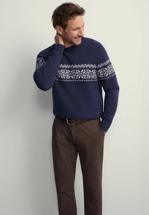 Granatowy sweter z białym geometrycznym wzorem na klatce piersiowej, w zestawieniu z brązowymi spodniami. Sweter ma okrągły dekolt i ściągacze na rękawach.