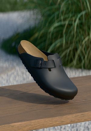 Schwarzer Schlupf-Clog mit seitlicher Schnalle und Korkfußbett, liegend auf einer Holzoberfläche mit grünem Laub im Hintergrund.