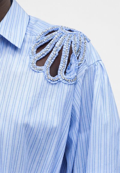 Φανέλα ανοιχτού γαλάζιου χρώματος με ρίγες και σχέδιο με κομμένη ράχη στον ώμο, περιγράμματα από ραμμένο ύφασμα και ασημένιες χάντρες διακόσμησης.