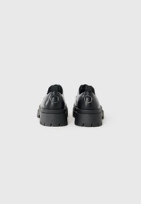 Mocasines de cuero negro grueso con suelas de goma gruesa y detalle de anillo metálico en las pestañas traseras, mostrados desde una vista trasera.