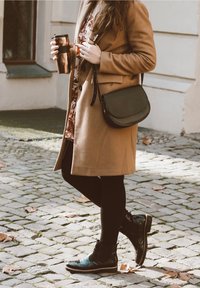 Cappotto di lana marrone, vestito floreale, borsa a tracolla nera, leggings neri e stivaletti neri lucidi, con in mano una tazza da viaggio in rame. Sfondo di ciottoli.