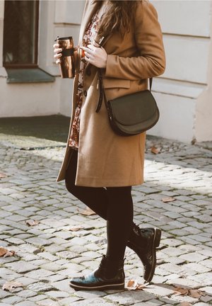 Cappotto di lana marrone, vestito floreale, borsa a tracolla nera, leggings neri e stivaletti neri lucidi, con in mano una tazza da viaggio in rame. Sfondo di ciottoli.