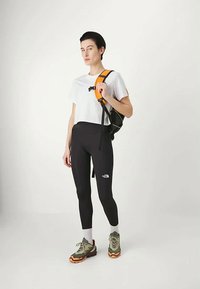 Nő fekete leggingsben, fehér pólóban, szürke és narancssárga túracipőben, feket és narancssárga hátizsák pántját a vállán viselve.