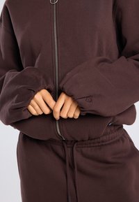 Brown zip-up jacket with voluminous sleeves and gathered cuffs, featuring a silver zipper and a minimal logo detail on the cuff.