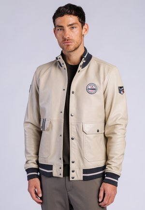 Mand iført beige bomberjakke med sort-hvid stribet krave, manchetter og kant, stående mod en ensartet lys baggrund.
