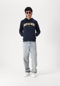 Marineblauwe hoodie met het "JACK & JONES" logo in het geel, gecombineerd met lichtblauwe jeans en witte sneakers. Model draagt zwarte zonnebril.