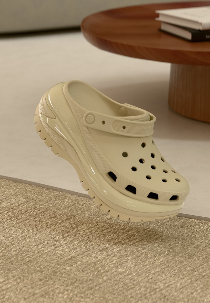 Sabot beige à enfiler avec des trous de ventilation et une semelle épaisse, placé sur un tapis beige texturé à côté d'une table ronde en bois avec des livres.