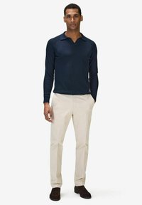 Mörkblå ribbad långärmad polo med V-ringning, matchad med krämfärgade byxor och mörkbruna skor. Modellen står upprätt.