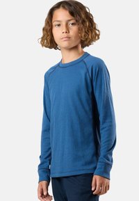 Young person with curly hair wearing a ribbed blue long-sleeve shirt and dark pants, standing against a plain light background.