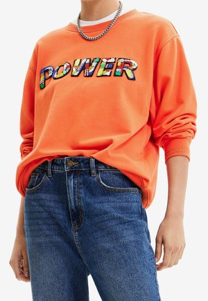 Persona che indossa una felpa arancione con scritta colorata "POWER" e jeans blu, accessoriata con una collana a catena d'argento.