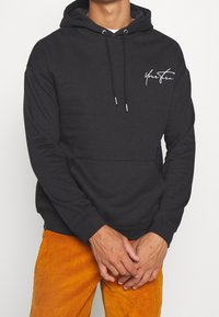 Sudadera con capucha negra hecha de una suave mezcla de algodón, que cuenta con un bolsillo frontal, capucha con cordón y un logo en cursiva blanca en el lado izquierdo del pecho.