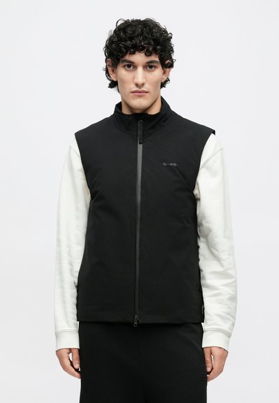 Fekete, ujjatlan mellény magas gallérral és elülső cipzárral. Simább anyagú textúrával és diszkrét logóval a mellrészen.