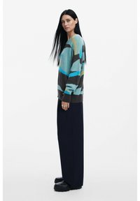 Maglione oversize colorato in turchese, marrone e grigio con motivi astratti, abbinato a pantaloni neri a gamba larga e scarpe nere chunky.
