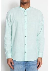 Camicia a maniche lunghe verde menta chiaro con colletto a fascetta, bottoni neri e tessuto testurizzato. Indossata con jeans azzurri chiari.