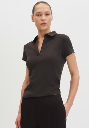 Femme portant une chemise noire à manches courtes, col et encolure en V avec un pantalon noir, debout, une main dans la poche, sur un fond uni.