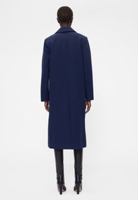 Hosszú tengerészkék kabát tűcsíkos mintával, hajtókás gallérral és egyenes szabással. Fekete magassarkú csizmával viselve. Hátulnézet.