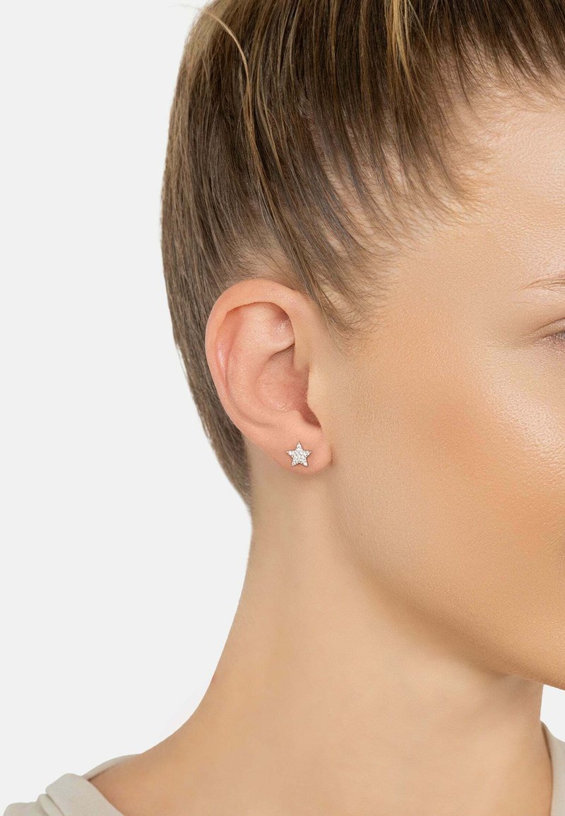 Primer plano de la oreja de una mujer que lleva un pequeño pendiente en forma de estrella de plata con detalles brillantes sobre un fondo sencillo.