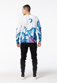 Camisa de manga larga blanca con patrones abstractos de olas en azul y rosa en las mangas, que presenta un gráfico de una persona practicando kitesurf. Pantalones y zapatos negros.