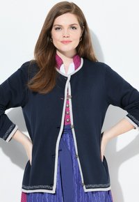 Cárdigan navy con bordes grises, cuello redondo, cierre de botones y adornos bordados en las mangas. Llevado sobre una blusa estampada y una falda.