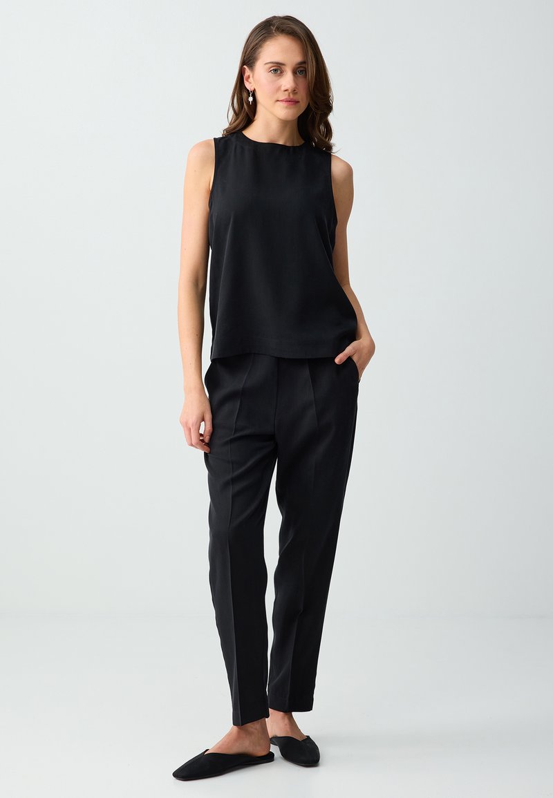 Femme portant un haut noir sans manches et un pantalon noir sur mesure avec des chaussures noires sans lacets, debout avec une main dans la poche devant un fond uni.